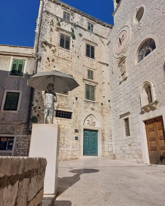 A bronze statue on a stone square beneath the old town clock in Šibenik